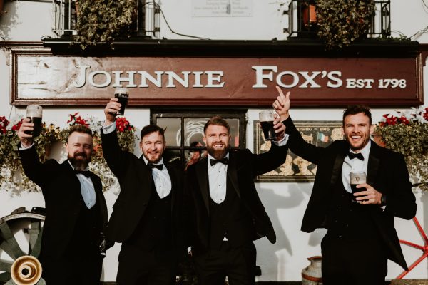 Groom and groomsmen raising pints of Guinness outside Johnnie Fox’s Pub in front of the iconic signage.