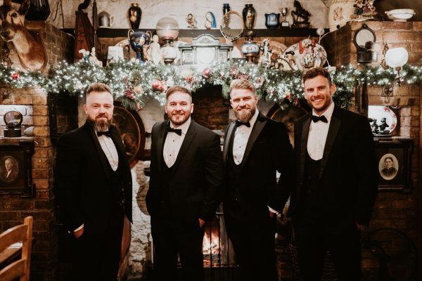 Groomsmen in tuxedos posing in front of a decorated fireplace at Johnnie Fox’s