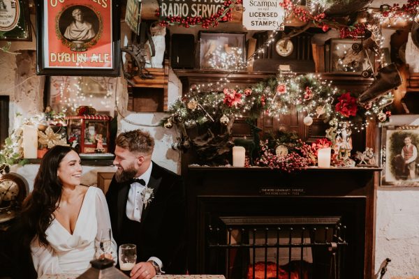 Bride and groom sitting together inside Johnnie Fox’s Pub, smiling beside a decorated fireplace with festive lights.