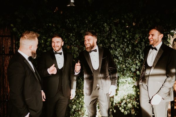 Groomsmen laughing together in tuxedos against ivy backdrop at Johnnie Fox’s