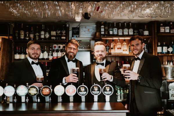 Groomsmen standing together behind the bar at Johnnie Fox’s, holding pints of Guinness during the wedding celebration.