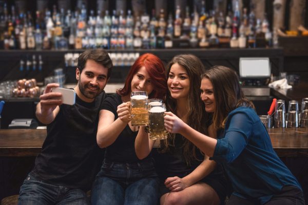 Friends holding beer glasses and taking a selfie at bar counter using mobile phone